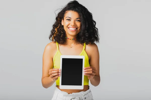 Mujer afroamericana feliz en la tapa amarilla de la cosecha que sostiene la tableta digital con la pantalla en blanco aislada en gris - foto de stock