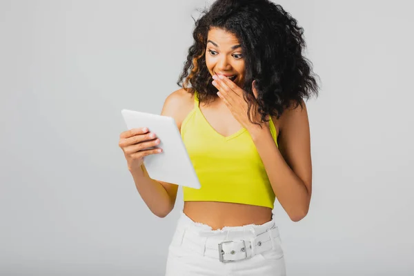 Surpris femme afro-américaine en haut de culture jaune regardant tablette numérique isolé sur gris — Photo de stock