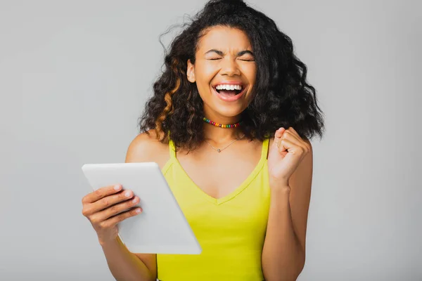 Alegre afroamericana mujer en amarillo recorte celebración tableta digital aislado en gris - foto de stock