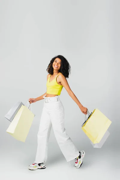 Pleine longueur de femme afro-américaine heureuse marchant tout en tenant des sacs à provisions sur gris — Photo de stock