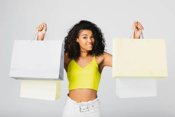 Cheerful african american woman holding shopping bags and biting lip isolated on grey — Stock Photo