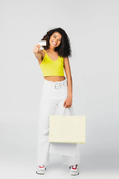 Mujer afroamericana feliz sosteniendo bolsas de papel y tarjeta de crédito en gris - foto de stock