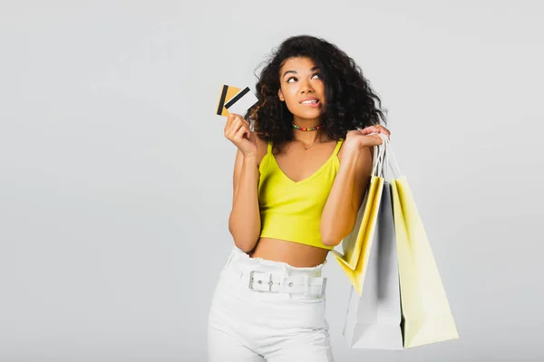 Femme afro-américaine réfléchie tenant des sacs en papier et des cartes de crédit isolés sur gris — Photo de stock