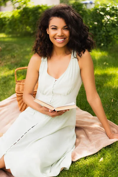 Gioiosa donna afro-americana in abito che tiene libro e seduta su una coperta nel parco — Foto stock