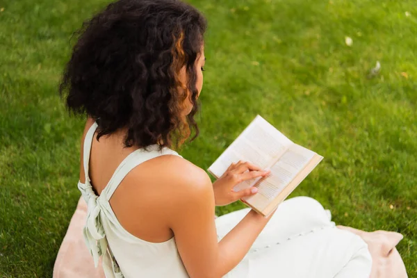 Curly african american woman in dress reading book and sitting on blanket on green lawn — Stock Photo
