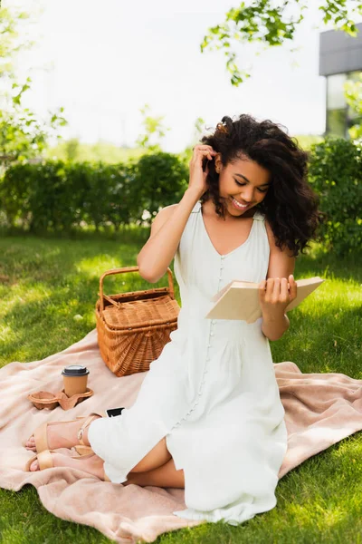 Femme afro-américaine gaie en robe lecture roman et assis sur la couverture dans le parc — Photo de stock