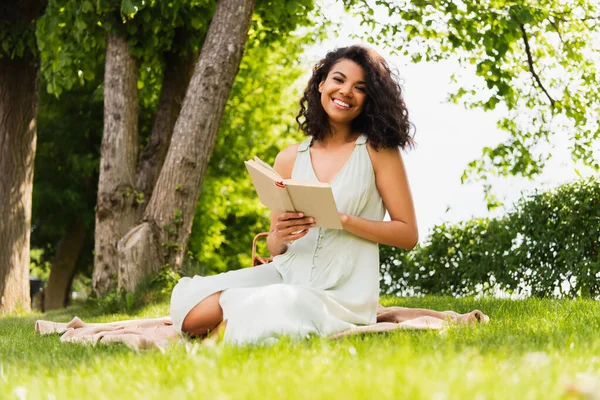 Alegre afroamericana mujer en vestido celebración libro y sentado en manta en parque - foto de stock