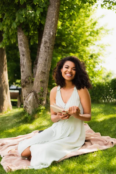 Happy african american woman in dress holding book and sitting on blanket in park — Stock Photo