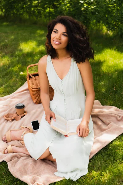 Femme afro-américaine rêveuse en tenue de robe livre pendant le pique-nique dans le parc — Photo de stock