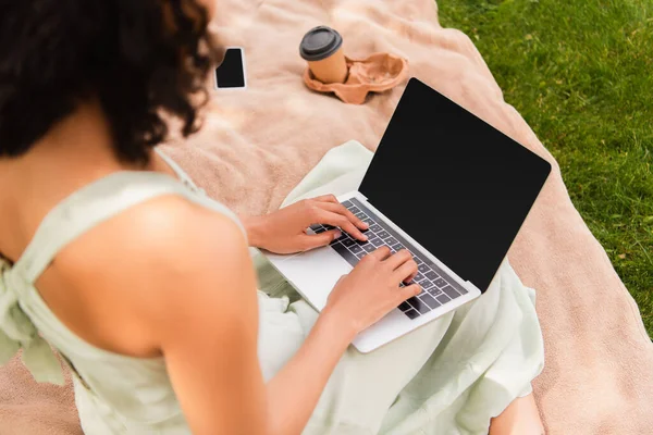 High angle view of african american woman using laptop near smartphone and paper cup on blanket in park — Stock Photo
