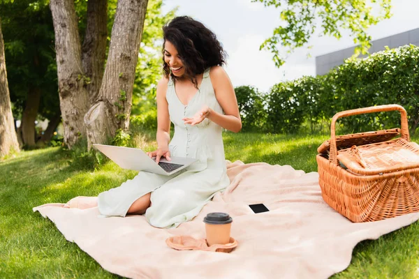 Mujer afroamericana positiva usando el ordenador portátil cerca de teléfono inteligente y canasta de mimbre en la manta en el parque - foto de stock