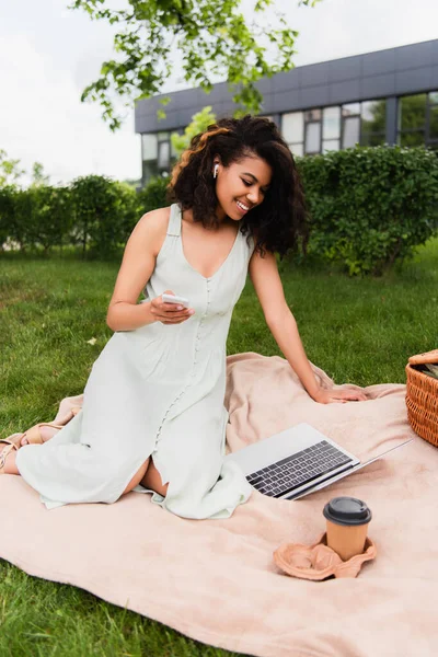Mujer afroamericana feliz usando el ordenador portátil y sosteniendo el teléfono inteligente cerca de la canasta de mimbre en la manta en el parque - foto de stock