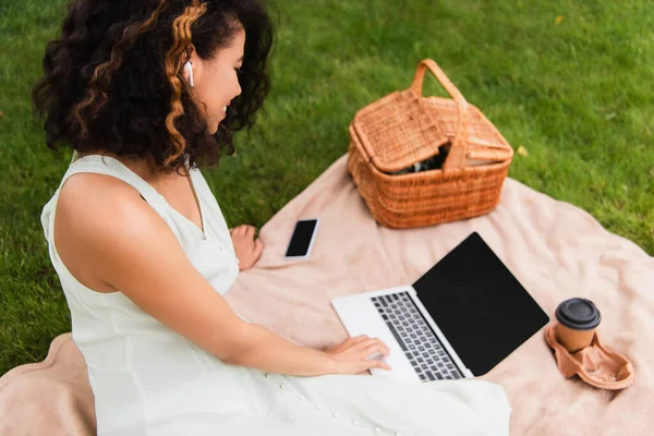 Encaracolado afro-americano mulher usando laptop perto cesta de vime e smartphone em cobertor no parque — Fotografia de Stock