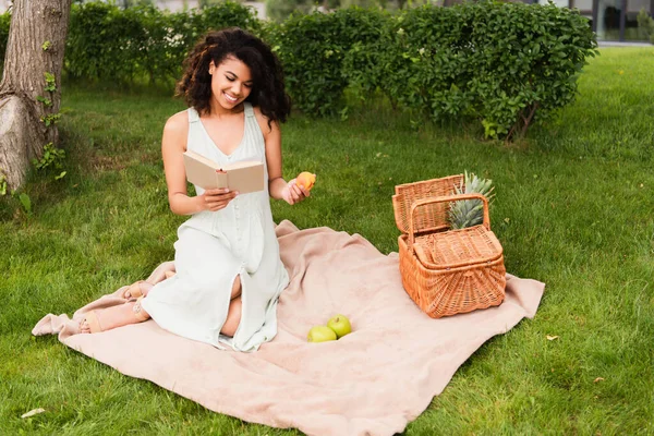 Smiling african american woman in white dress holding peach and reading book during picnic — Stock Photo