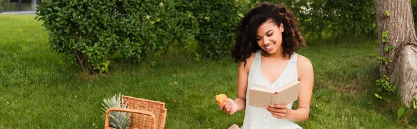 Mujer afroamericana feliz en vestido blanco sosteniendo melocotón y libro de lectura durante el picnic, pancarta - foto de stock