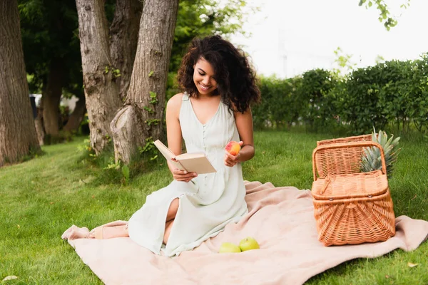 Happy african american woman in white dress holding peach and reading book during picnic — Stock Photo