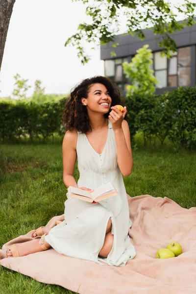Mujer afroamericana feliz en vestido blanco sosteniendo melocotón y libro durante el picnic - foto de stock