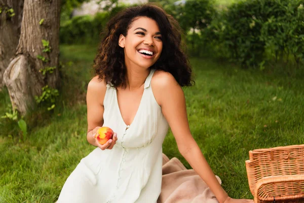 Mulher americana africana alegre em vestido branco segurando pêssego perto cesta de vime no parque — Fotografia de Stock