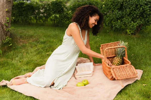 Mujer afroamericana feliz en vestido blanco sentado cerca de la cesta de picnic y frutas en manta - foto de stock