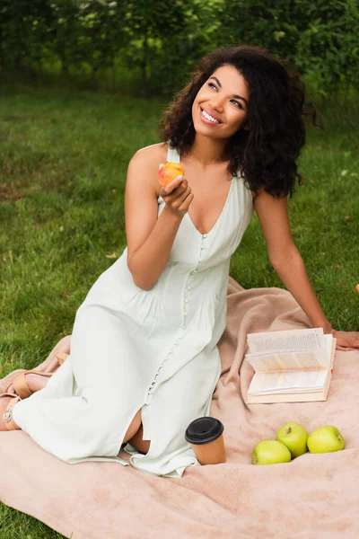 Happy and young african american woman in white dress holding peach in park — Stock Photo