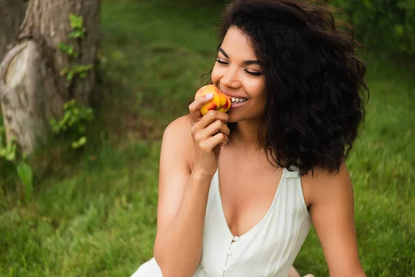 Happy and young african american woman in white dress eating peach in park — Stock Photo