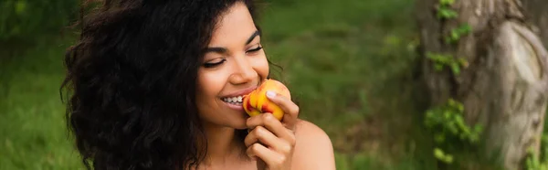 Glücklich und junge afrikanisch-amerikanische Frau isst Pfirsich im Park, Banner — Stockfoto