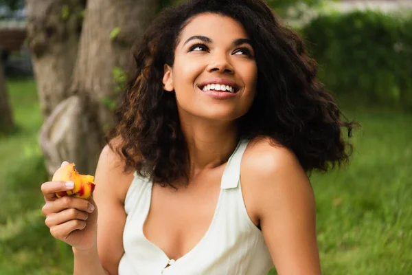 Счастливая и молодая африканская американка в белом платье держит персик в парке — стоковое фото