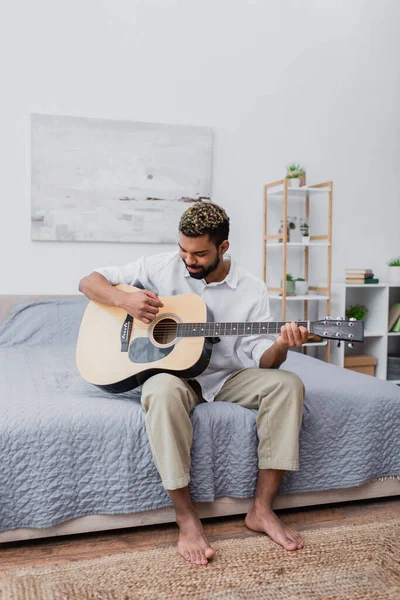 Feliz joven afroamericano hombre con el pelo teñido y la barba tocando la guitarra acústica en el dormitorio - foto de stock