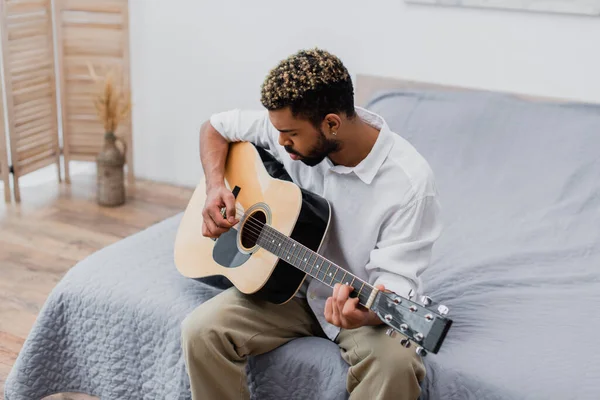 Barbudo joven afroamericano hombre con el pelo teñido tocando la guitarra acústica en el dormitorio - foto de stock