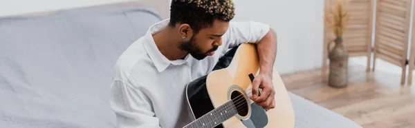 Barbuto giovane uomo afroamericano con i capelli tinti suonare la chitarra acustica in camera da letto, banner — Foto stock