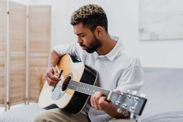 Fokussierter junger afrikanisch-amerikanischer Mann mit gefärbten Haaren spielt Akustikgitarre im Schlafzimmer — Stockfoto