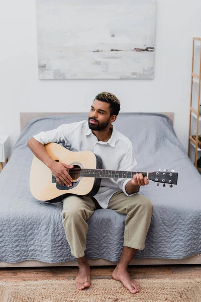 Joven afroamericano hombre con el pelo teñido tocando la guitarra acústica en dormitorio moderno - foto de stock
