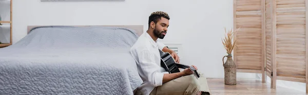 Giovane uomo afroamericano con i capelli tinti suonare la chitarra acustica vicino al letto, banner — Foto stock