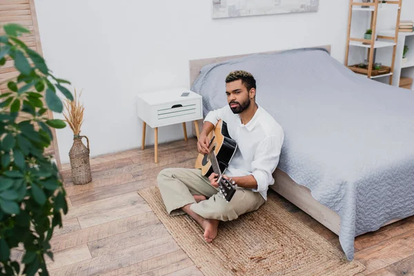 Homme afro-américain barbu avec les cheveux teints assis sur le tapis et jouer de la guitare acoustique près du lit — Photo de stock