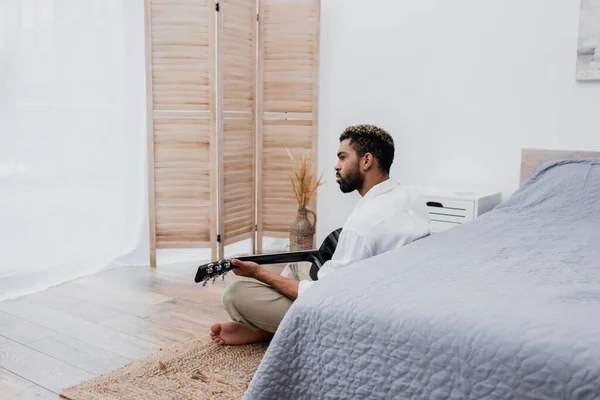 Homme afro-américain barbu assis sur le tapis et jouant de la guitare acoustique près du lit — Photo de stock