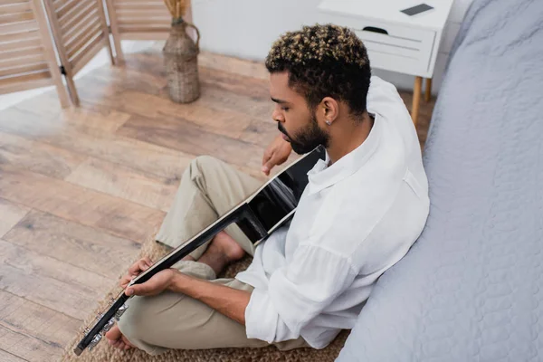 Vista de ángulo alto de barbudo joven afroamericano hombre con el pelo teñido tocando guitarra acústica cerca de la cama - foto de stock