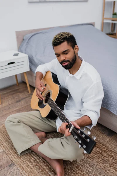 Barbudo hombre afroamericano sentado en la alfombra y tocando la guitarra acústica cerca de la cama y la mesita de noche - foto de stock