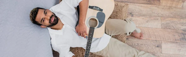 Vista superior do homem americano africano satisfeito com cabelo tingido segurando guitarra acústica e inclinando-se na cama, bandeira — Fotografia de Stock