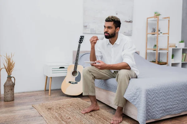 Молодой африканский американец с окрашенными волосами, держа чашку кофе и блюдце, сидя на кровати рядом с акустической гитарой — стоковое фото