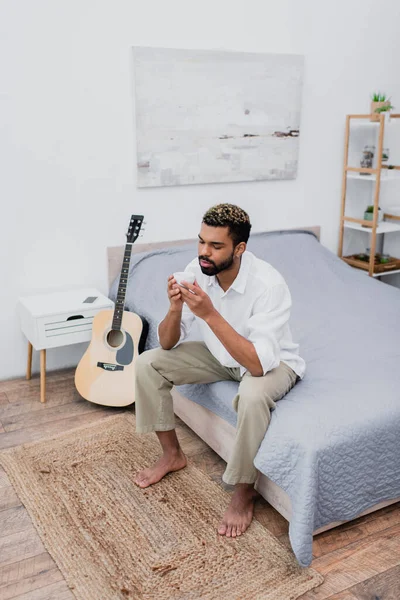 Vista ad alto angolo di uomo afro-americano in possesso di una tazza di caffè mentre seduto sul letto vicino alla chitarra acustica — Foto stock