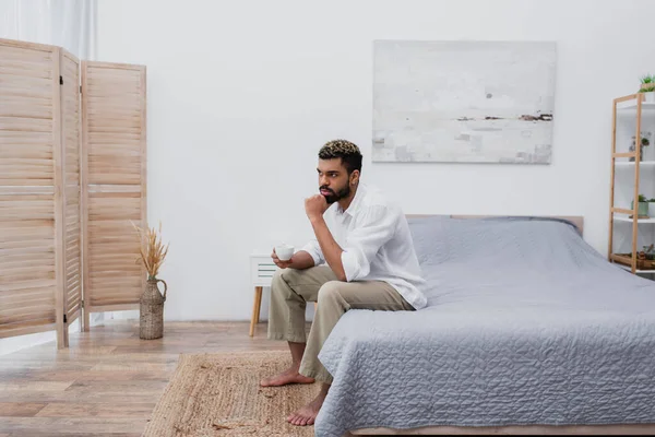 Nachdenklicher afrikanisch-amerikanischer Mann mit gefärbten Haaren, der eine Tasse Kaffee hält, während er zu Hause auf dem Bett sitzt — Stockfoto