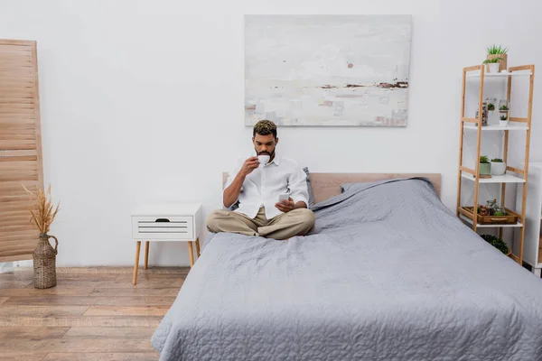 Junger afrikanisch-amerikanischer Mann mit gefärbten Haaren hält eine Tasse Kaffee in der Hand und benutzt das Smartphone, während er sich auf dem Bett ausruht — Stockfoto