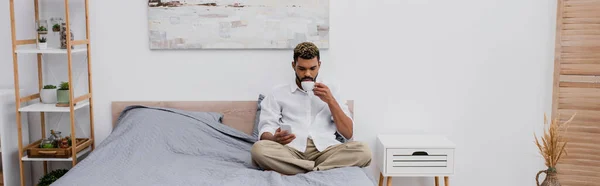 Young african american man holding cup of coffee and using smartphone while resting on bed, banner — Stock Photo