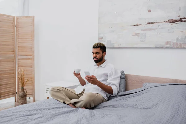 Bärtiger afrikanisch-amerikanischer Mann mit gefärbten Haaren, der eine Tasse Kaffee in der Hand hält und das Smartphone benutzt, während er sich im Bett ausruht — Stockfoto