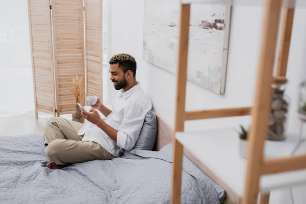Glücklicher afrikanisch-amerikanischer Mann mit gefärbten Haaren, der eine Tasse Kaffee in der Hand hält und das Smartphone benutzt, während er sich im Bett ausruht — Stockfoto