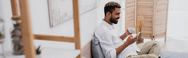 Heureux homme afro-américain avec les cheveux teints tenant tasse de café et en utilisant smartphone tout en se reposant sur le lit, bannière — Photo de stock