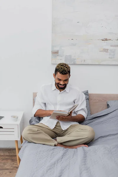 Happy african american man with dyed hair reading book in modern bedroom — Stock Photo