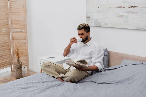 Giovane uomo afroamericano con libro di lettura dei capelli tinti e bere caffè in camera da letto moderna — Foto stock
