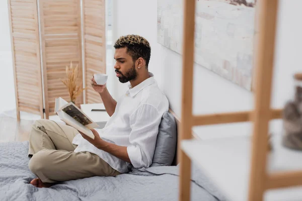 Junger afrikanisch-amerikanischer Mann mit gefärbten Haaren liest Buch und hält Tasse Kaffee im modernen Schlafzimmer — Stockfoto