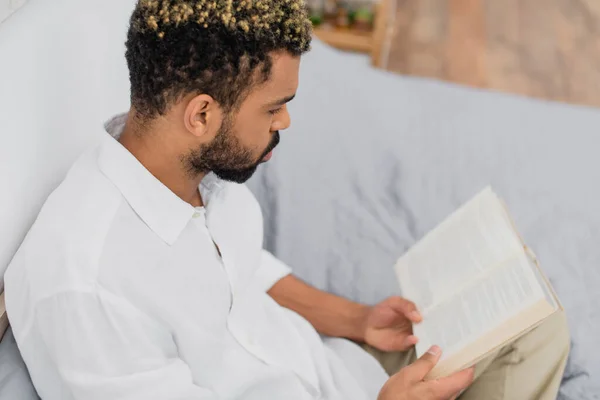 Vista ad alto angolo di giovane uomo afroamericano con capelli tinti lettura libro in camera da letto — Foto stock